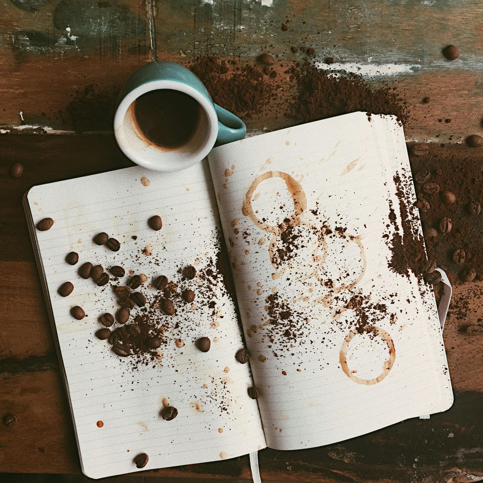 Overhead view of coffee-stained notepad and scattered beans with a mug on a rustic wooden table.